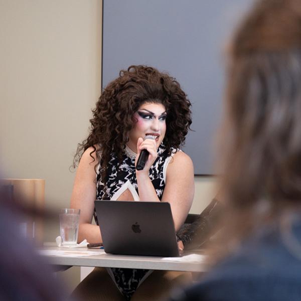A performer with curly hair speaks into a microphone at a table with a laptop and a glass of water.