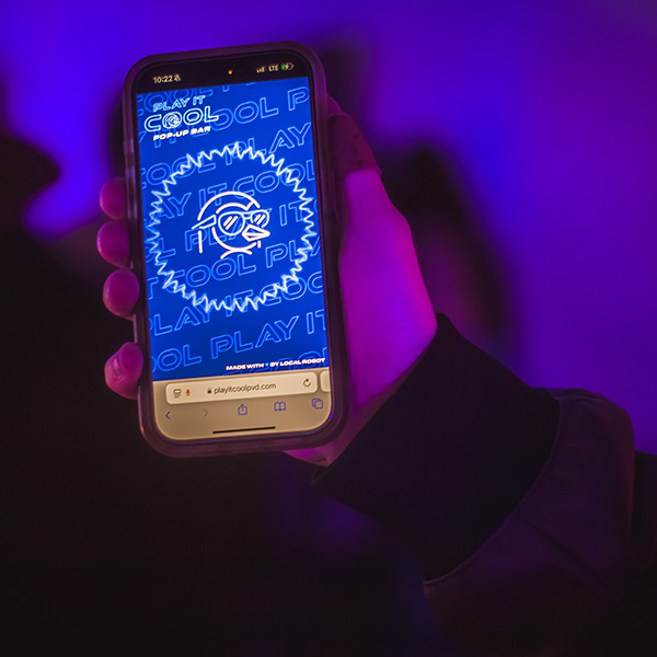 A hand holds a smartphone displaying a blue graphic for a pop-up bar event.