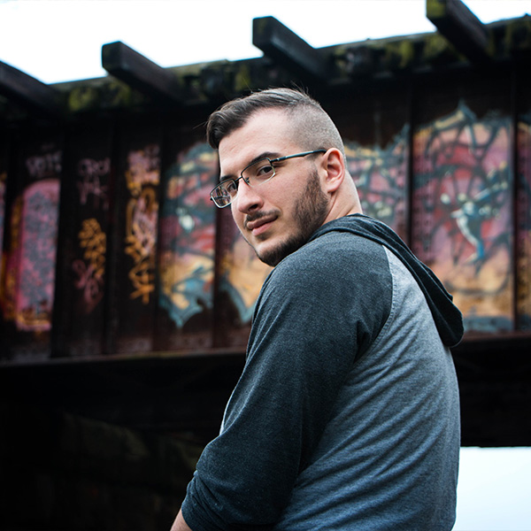A young man with glasses looks back, standing under a graffiti-covered structure.