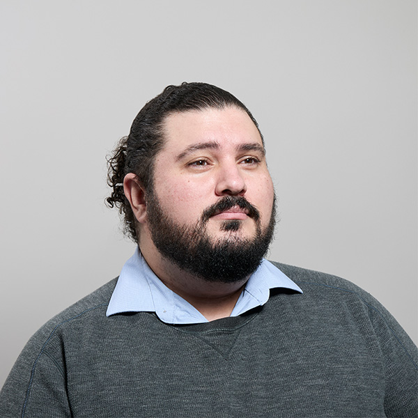 A man with a beard and curly hair looks thoughtfully to the side, wearing a gray sweater and blue shirt.