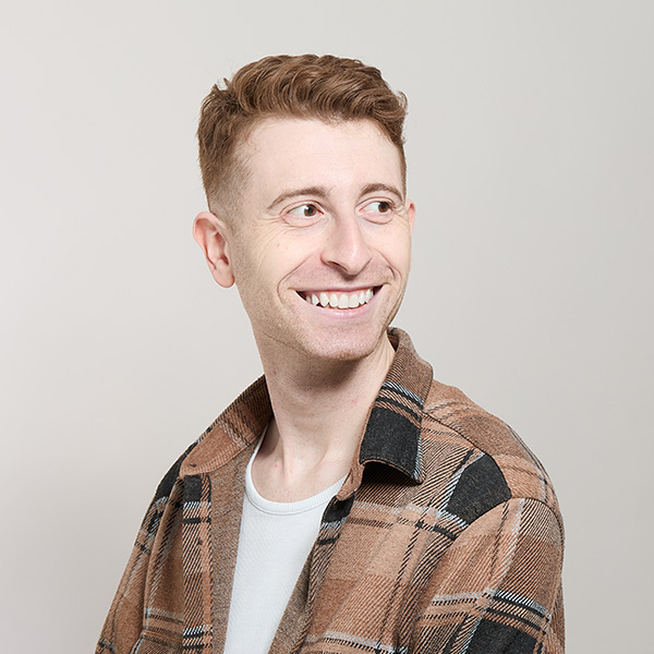 A smiling person with short, light brown hair looks to the side, wearing a plaid shirt over a white shirt.